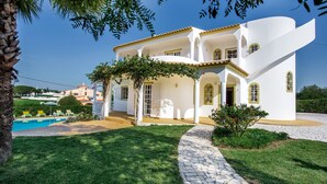 A white villa with a stone pathway, a swimming pool, and a lush garden.