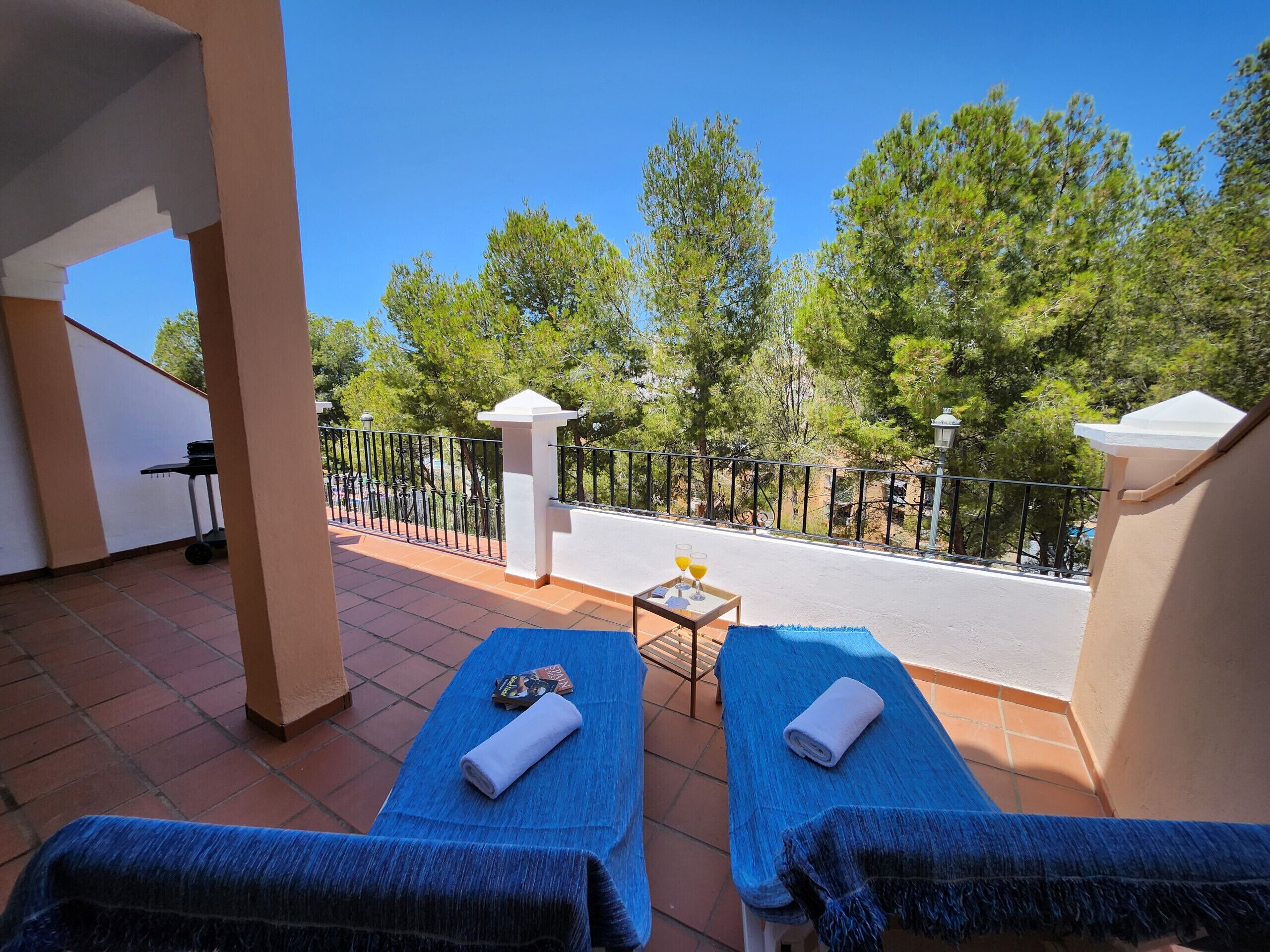 A balcony with two sunbeds, a small table with drinks, and a view of trees and a road.