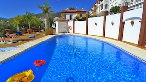 A swimming pool with clear blue water, surrounded by a white wall with brown accents, and a view of a residential area with houses and palm trees in the background. - 3 Bedrooms | Aljamar 19A | CG  R1086 (Nerja)