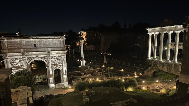 An ancient Roman site at night with illuminated ruins, including an archway and columns.