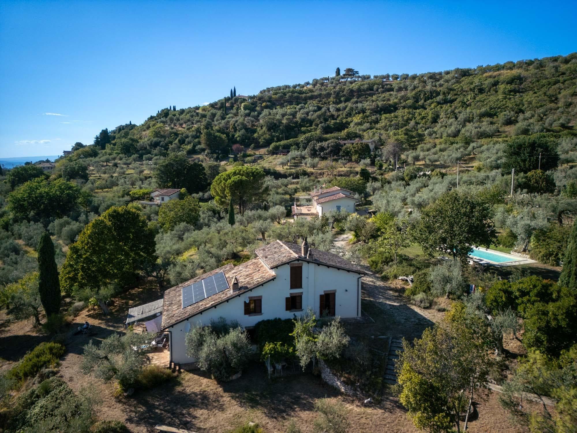 A rural house with solar panels, surrounded by trees and a hill in the background.