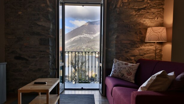 A room with a stone wall, a red sofa, a wooden table, and a view of a mountain and lake through an open door.