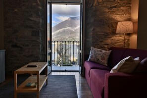 Une pièce avec un mur en pierre, un canapé rouge, une table en bois et une vue sur une montagne et un lac, visible à travers une porte ouverte.