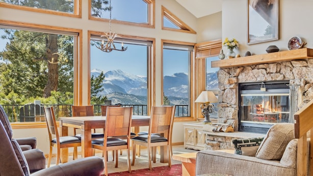 A cozy living room with a stone fireplace, large windows offering mountain views, and a dining area with wooden furniture. - Dances With Wolves Vacation At Windcliff 3 Bedroom Home by RedAwning (Estes Park)