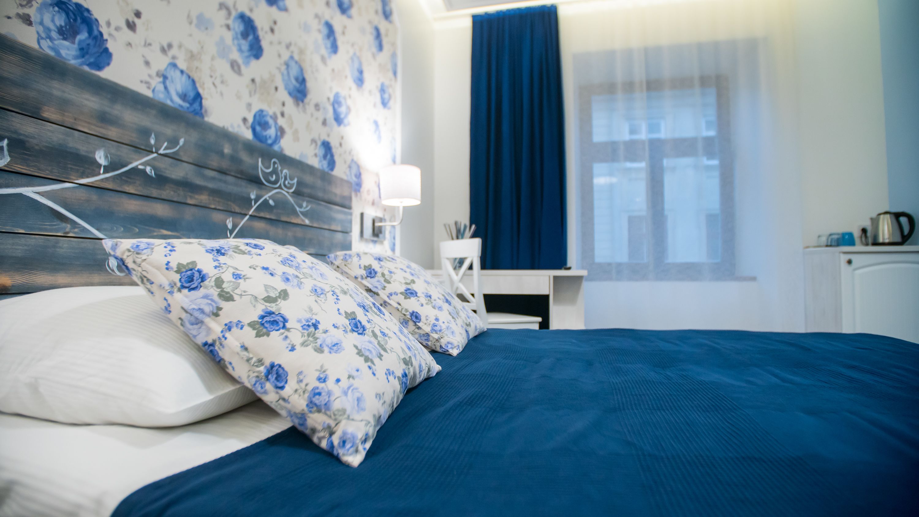 A bedroom with a bed, floral-patterned pillows, a wooden headboard, a nightstand with a lamp, and a window with curtains.