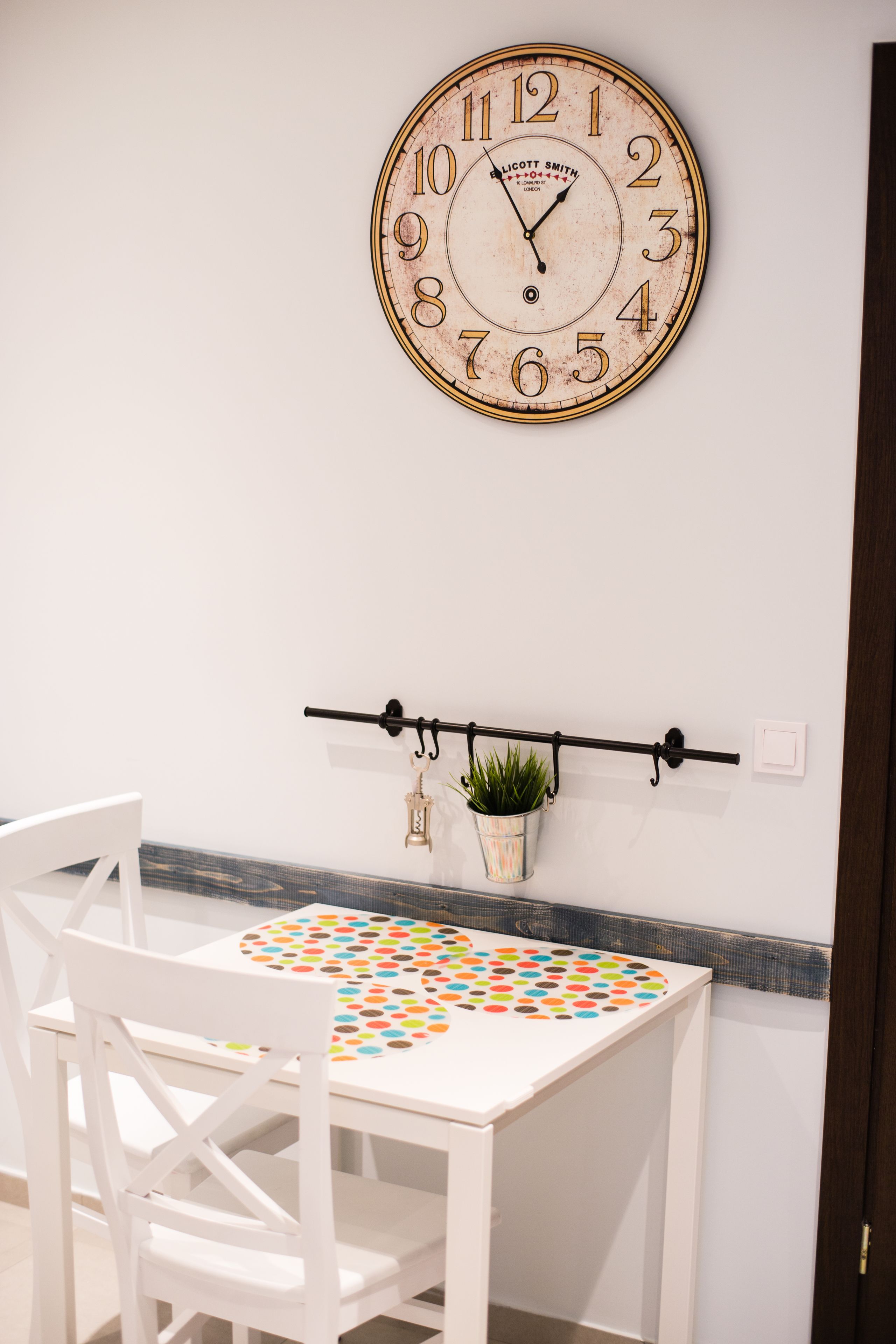 A white dining table with a colorful circular table runner, a white chair, and a wall-mounted clock.