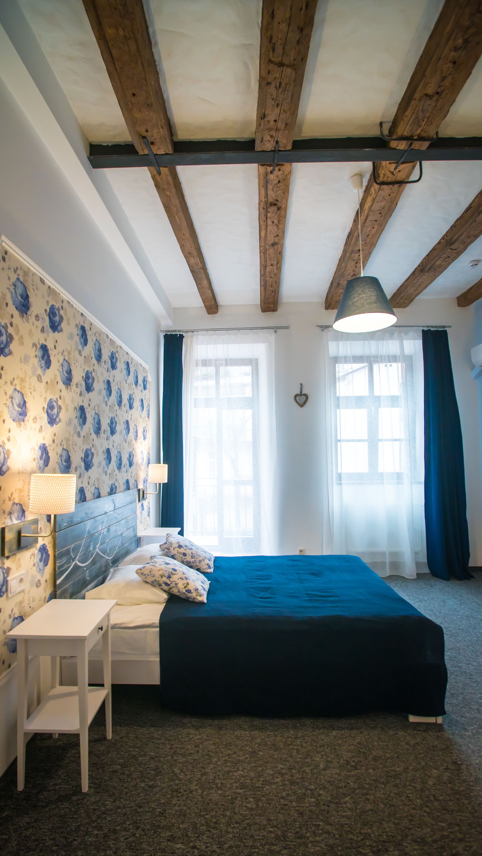 Une chambre avec un plafond en bois, un lit recouvert d’une courtepointe bleue, une table de chevet blanche, une lampe et un mur recouvert d’un papier peint à motifs floraux.