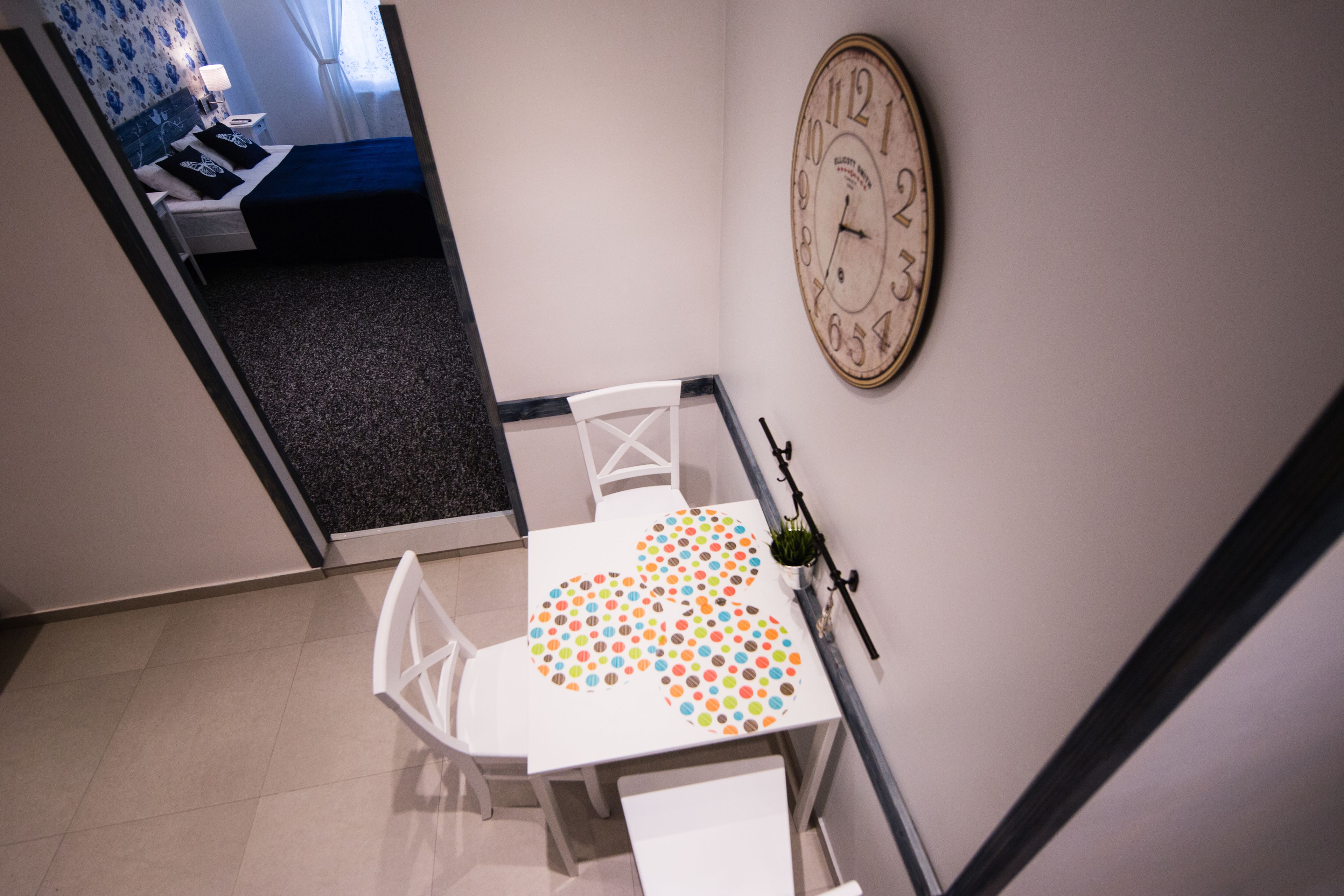 Un coin repas avec une table et des chaises blanches, une horloge au mur et une vue sur une autre pièce.