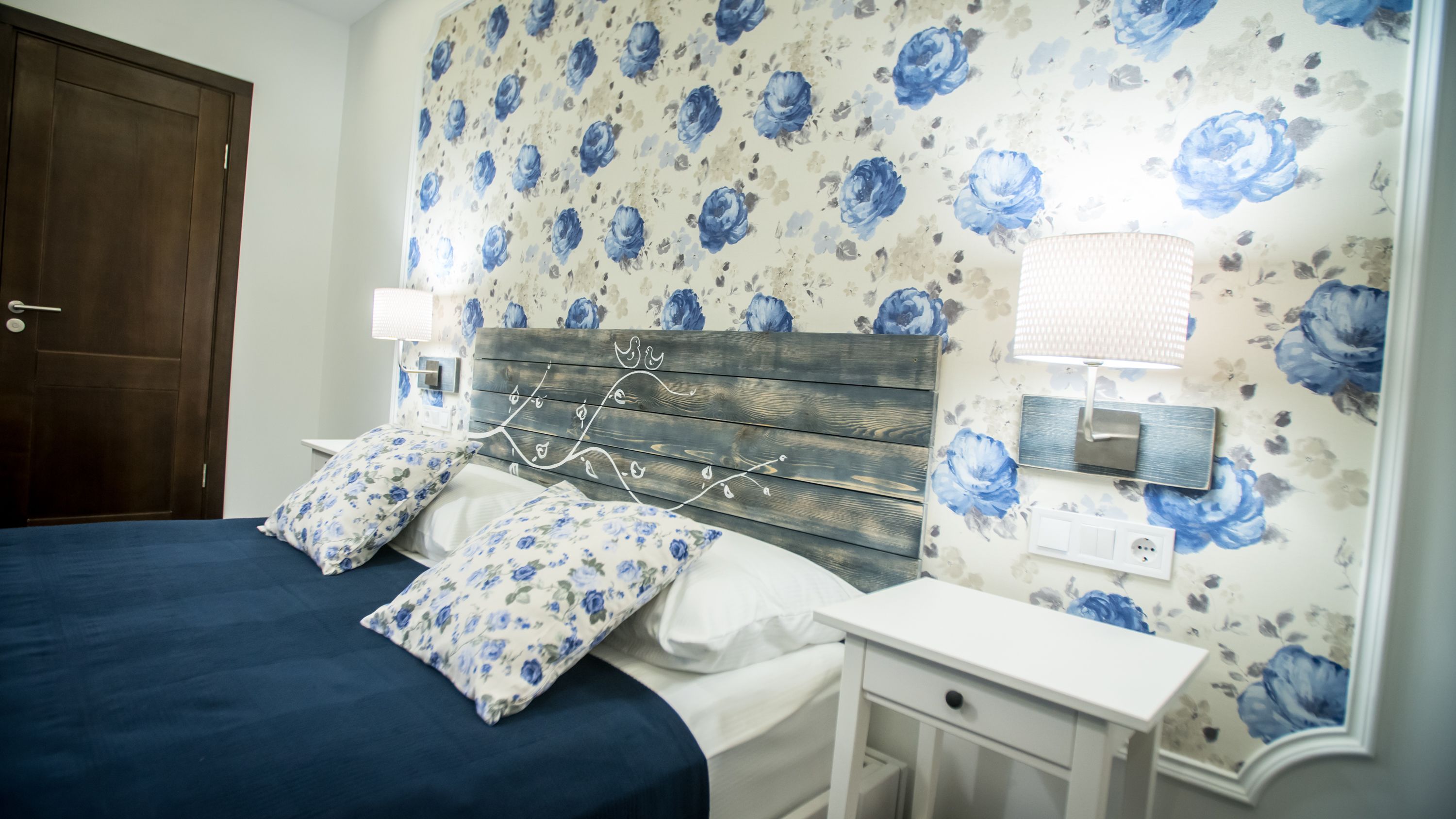A bedroom with a bed, two nightstands, a wooden headboard, floral wallpaper, and a door.