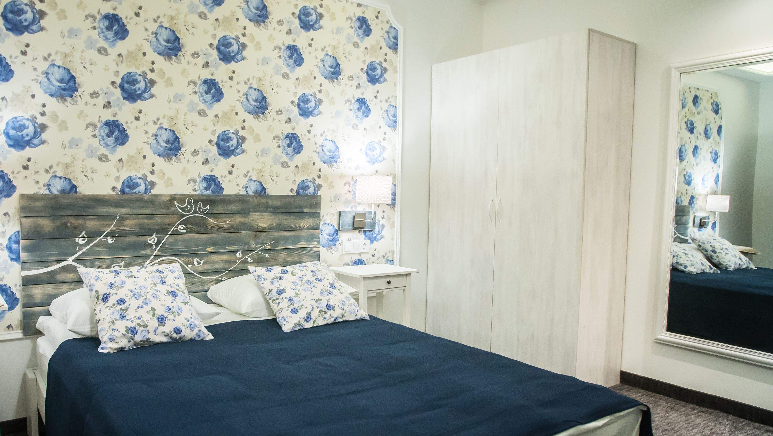 A bedroom with a floral wallpaper, a wooden headboard, a bed with blue bedding, a nightstand, a mirror, and a wardrobe.