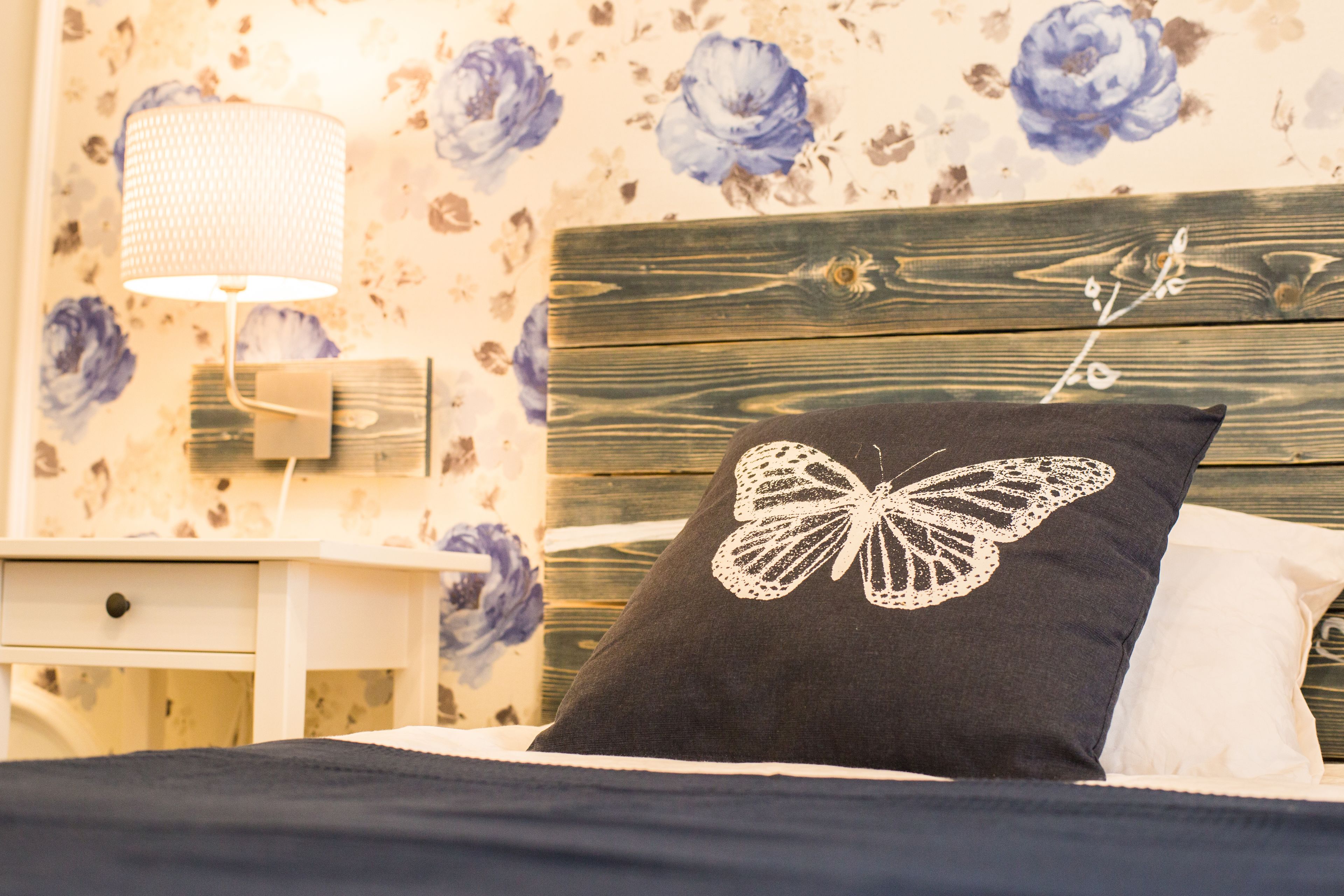 A bedroom with a wooden headboard, a black pillow with a white butterfly design, a white bedside lamp, and floral wallpaper.