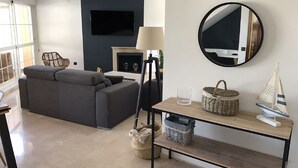 A modern living room with a dark-colored sofa, a flat-screen TV mounted on the wall, a fireplace, a round mirror, and a wooden console table with baskets and a model sailboat. - Beachfront Newly Renovated Duplex with Sea Views (Marbella)