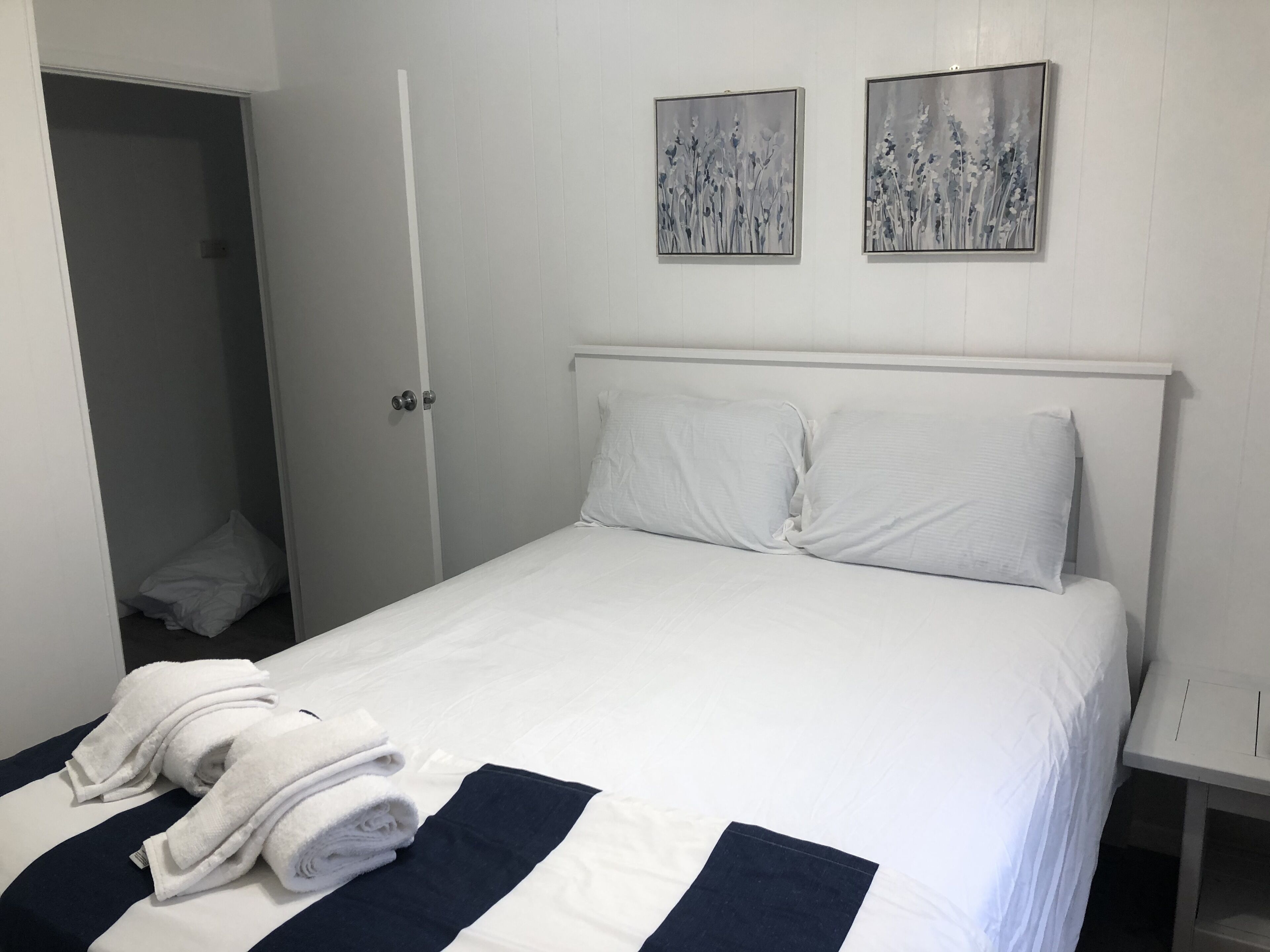 Een net opgemaakt bed met witte lakens en een marineblauwe accentkleur, een paar gevouwen handdoeken en twee ingelijste bloemschilderijen aan de muur.