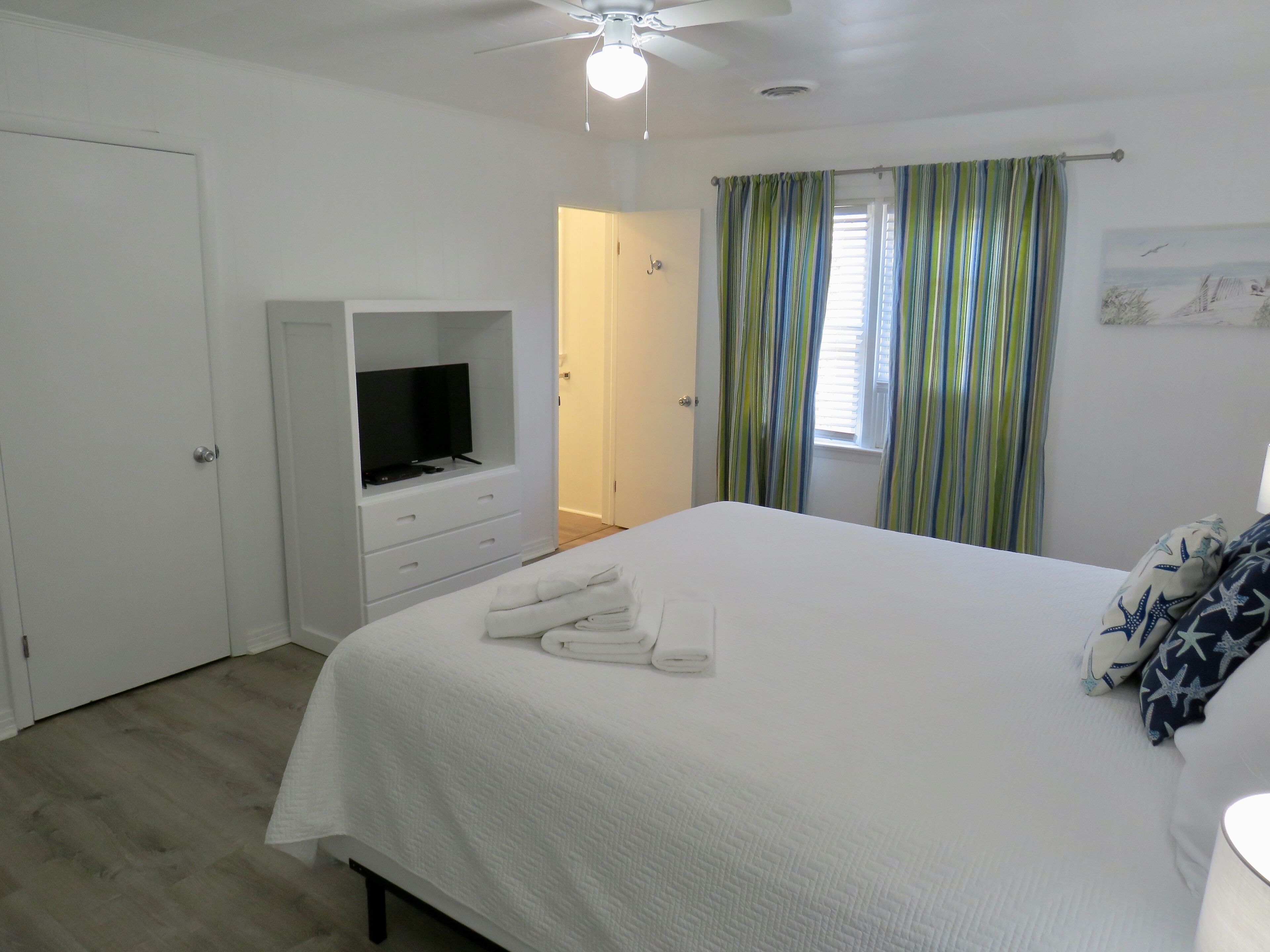 A bedroom with a bed, a TV in a built-in cabinet, a ceiling fan, and a window with curtains.