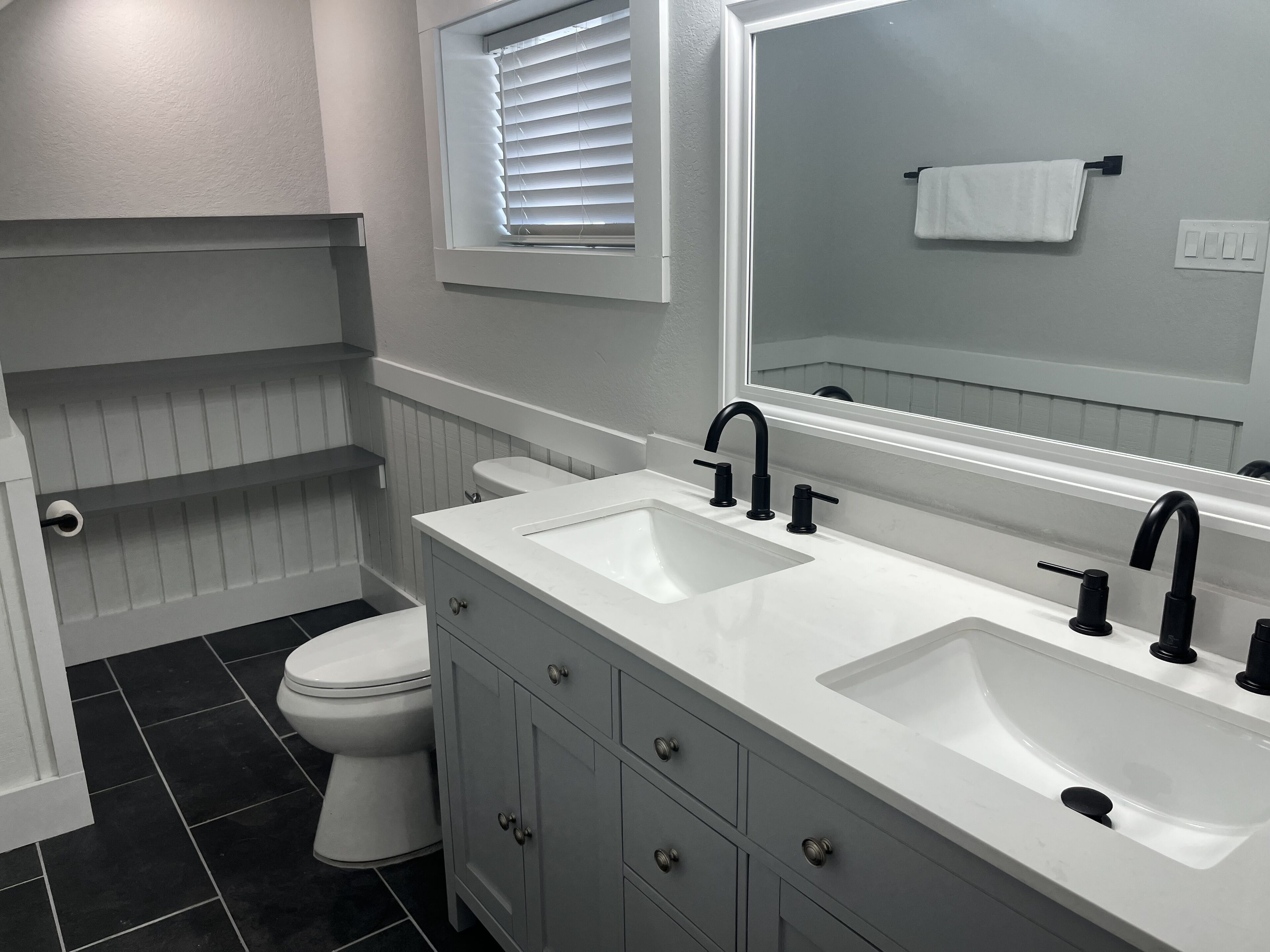 A bathroom with a white vanity, two sinks, a toilet, and a window with blinds.