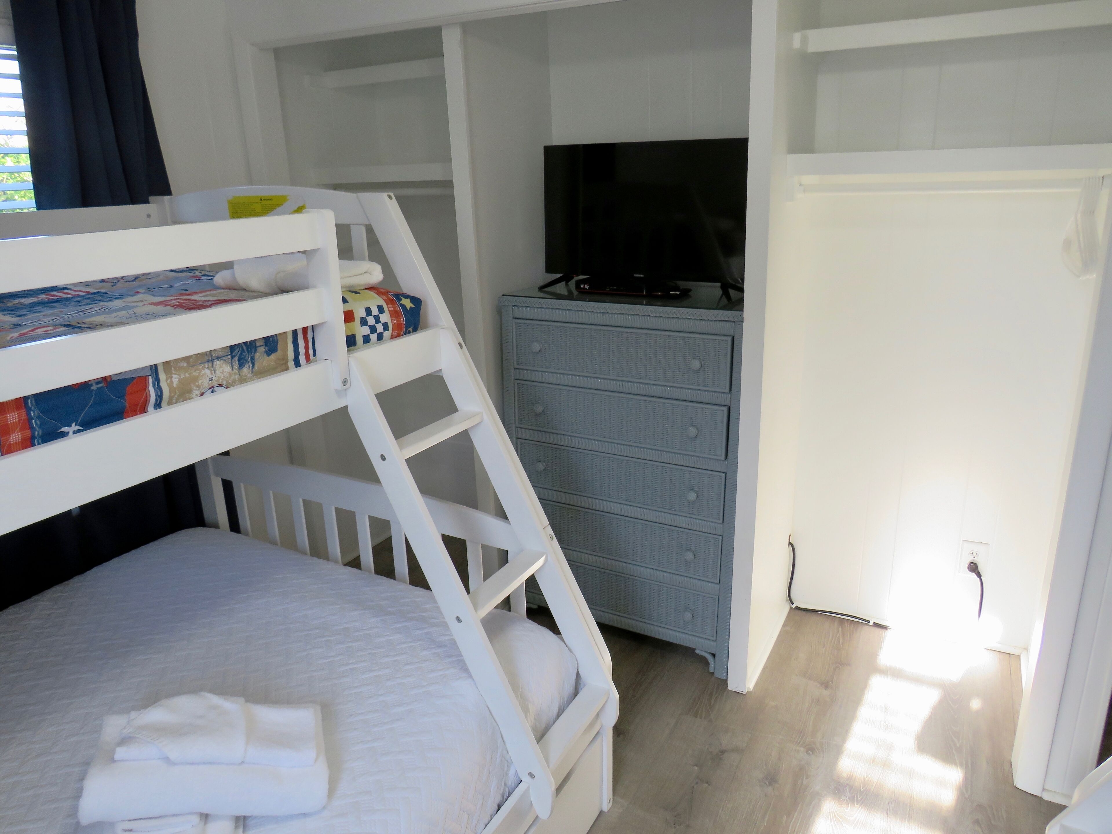 Un lit superposé avec une tête de lit matelassée, un petit bureau avec une télévision et une commode avec des tiroirs.