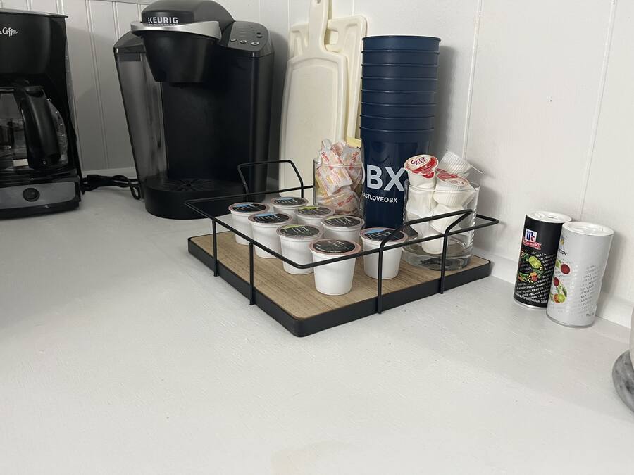 Una estación de café con una cafetera Keurig, una pila de vasos de papel, un dispensador de agua BX y un recipiente con cápsulas de café.