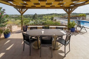 Un patio al aire libre con una mesa redonda y sillas, rodeado de plantas en macetas, con vistas a una piscina y un paisaje pintoresco.
