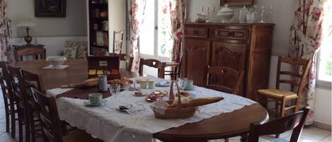 Une salle à manger avec une table ronde dressée pour six personnes, entourée de chaises en bois. On y trouve une bibliothèque, un meuble contenant de la verrerie et un tableau accroché au mur.