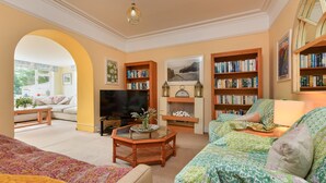 A cozy living room with a sofa, a coffee table, and a TV. There are bookshelves filled with books and a fireplace with a clock above it. - GORGEOUS SPACIOUS  FAMILY HOUSE IN THE CENTRE OF ABERSOCH (Abersoch)