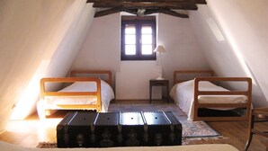 Une petite pièce aménagée comme une chambre sous les toits, avec deux lits en bois, une fenêtre, une lampe et un coffre.