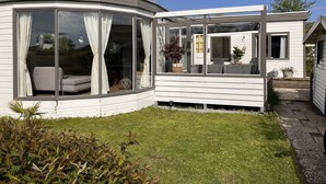 Une maison mobile moderne, de couleur blanche, dotée de grandes fenêtres en verre, d’une façade incurvée et d’un petit jardin.