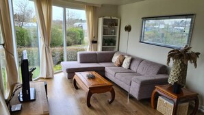Un salon avec un canapé gris, une table basse en bois, un téléviseur posé sur un meuble en bois, un vase décoratif et une fenêtre avec des stores.