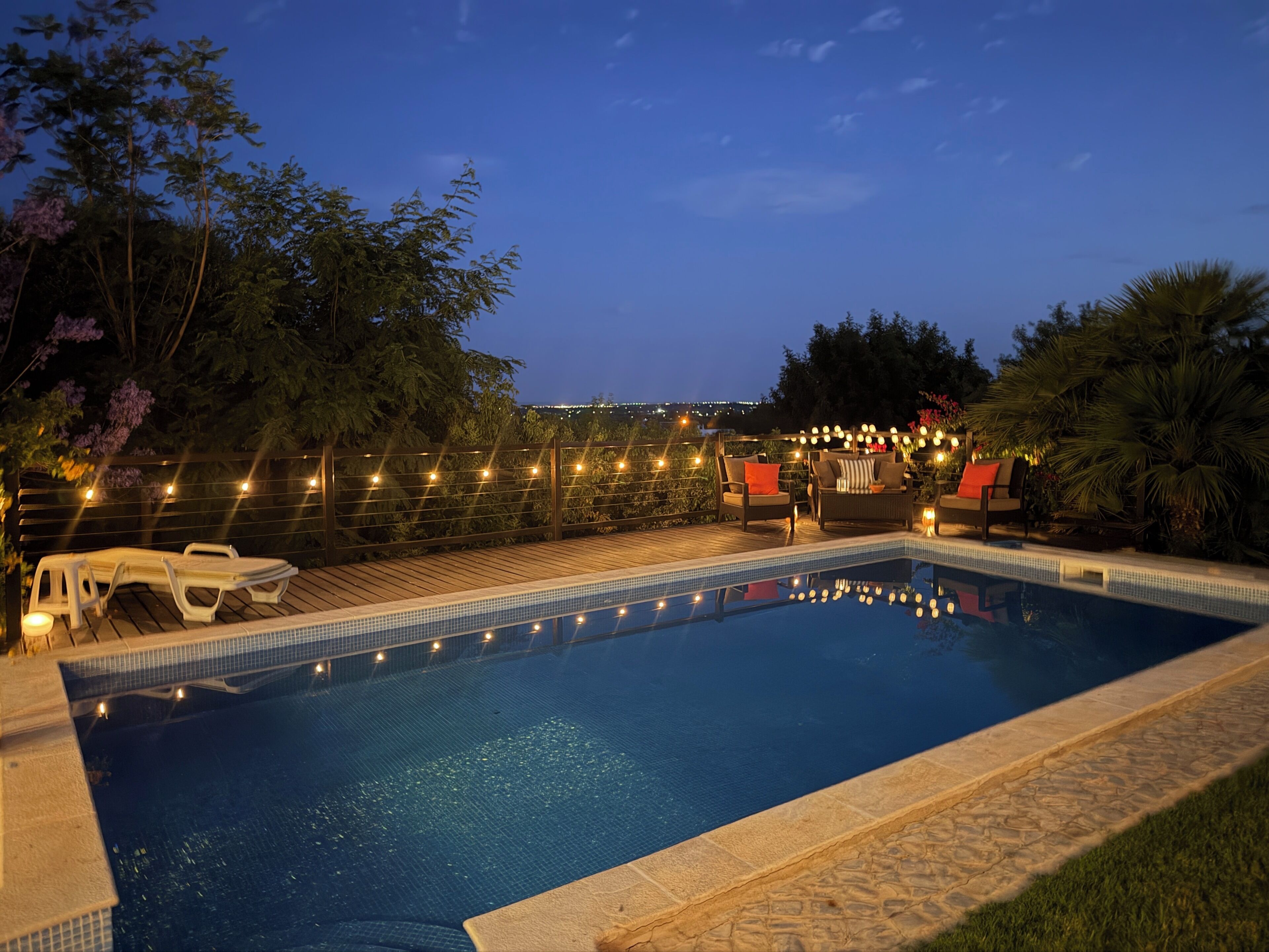 Una piscina rectangular con borde de azulejos, rodeada por una terraza de madera y una guirnalda de luces a lo largo de la valla.