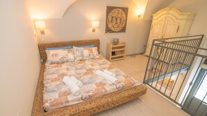Un dormitorio con una cama de mimbre, dos lámparas de pared, un cuadro, un armario de madera y la barandilla de un balcón.