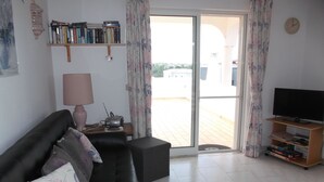 A living room with a black leather sofa, a television on a wooden stand, a bookshelf with books, a lamp, and a sliding glass door leading outside. - Holiday apartment ideal for a couple with private Swimming pool (Carvoeiro)