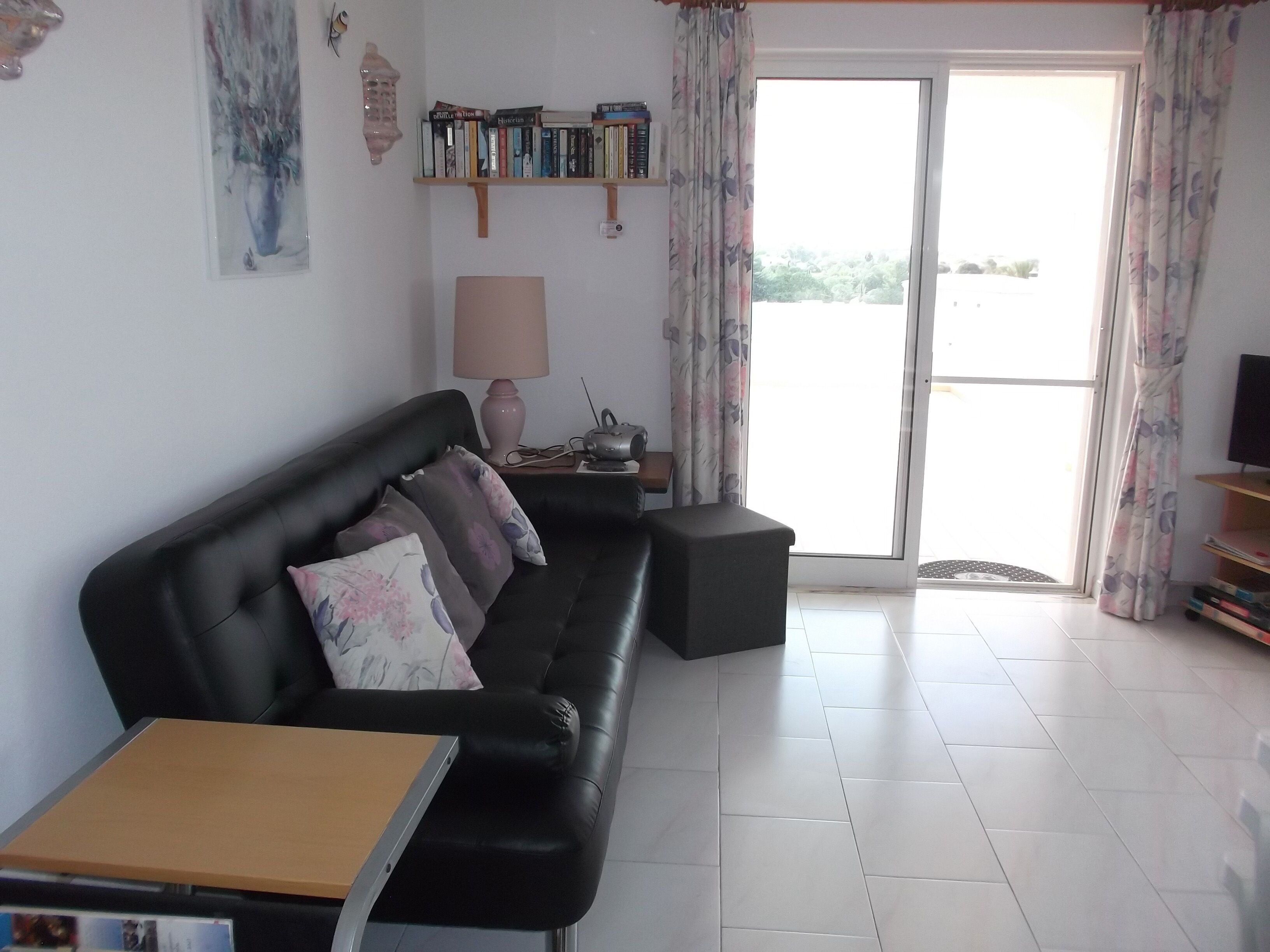 A living room with a black leather sofa, a small wooden table, a lamp, a bookshelf, and a sliding glass door leading to an outdoor area.
