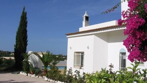 A white building with a tiled roof, a chimney, and a window with bars. There is a tall tree and a bush with pink flowers in the foreground. - Holiday apartment ideal for a couple with private Swimming pool (Carvoeiro)