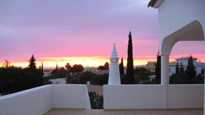 A balcony with two white chairs, a small table, and a view of a white tower and buildings against a sunset sky. - Holiday apartment ideal for a couple with private Swimming pool (Carvoeiro)