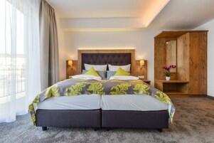 Una cama doble con cabecera capitonada, dos mesitas de noche con lámparas, un armario de madera y un ventanal con cortinas translúcidas.