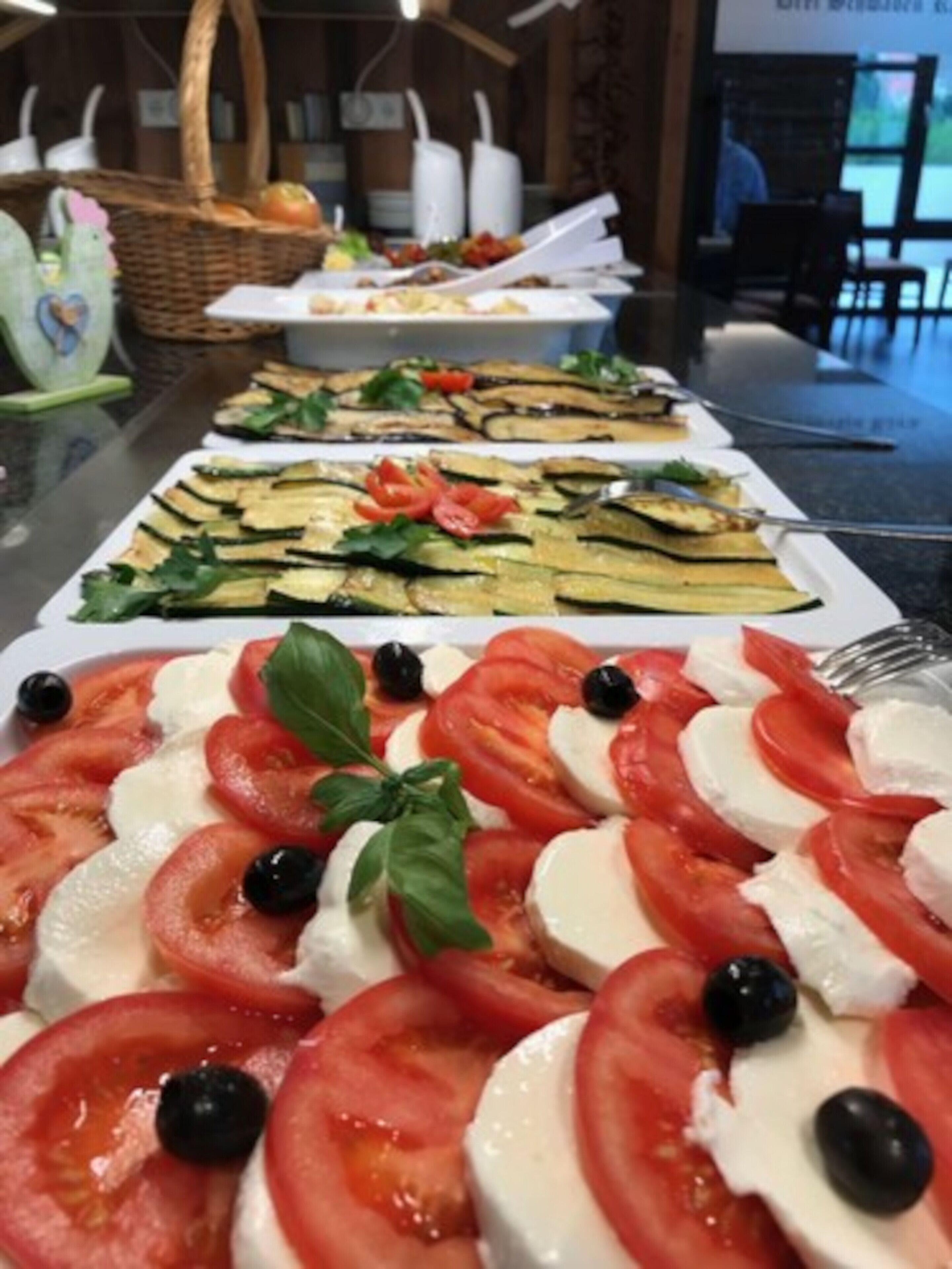 Uma mesa de buffet com diversos pratos, incluindo uma salada de tomate e mussarela, legumes grelhados e outras opções de comida pronta.