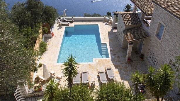 Une piscine rectangulaire bordée d’une terrasse en pierre, entourée de chaises longues et de plantes en pot, et offrant une vue sur une étendue d’eau.