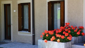 Un edificio con dos ventanas, cada una con contraventanas de madera, y una jardinera de hormigón con flores rojas al frente.