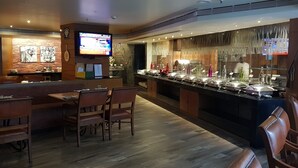 A restaurant with a long counter, wooden chairs, and a television screen displaying a program. - Regenta Place Bangalore, Cunningham Road (Bengaluru)