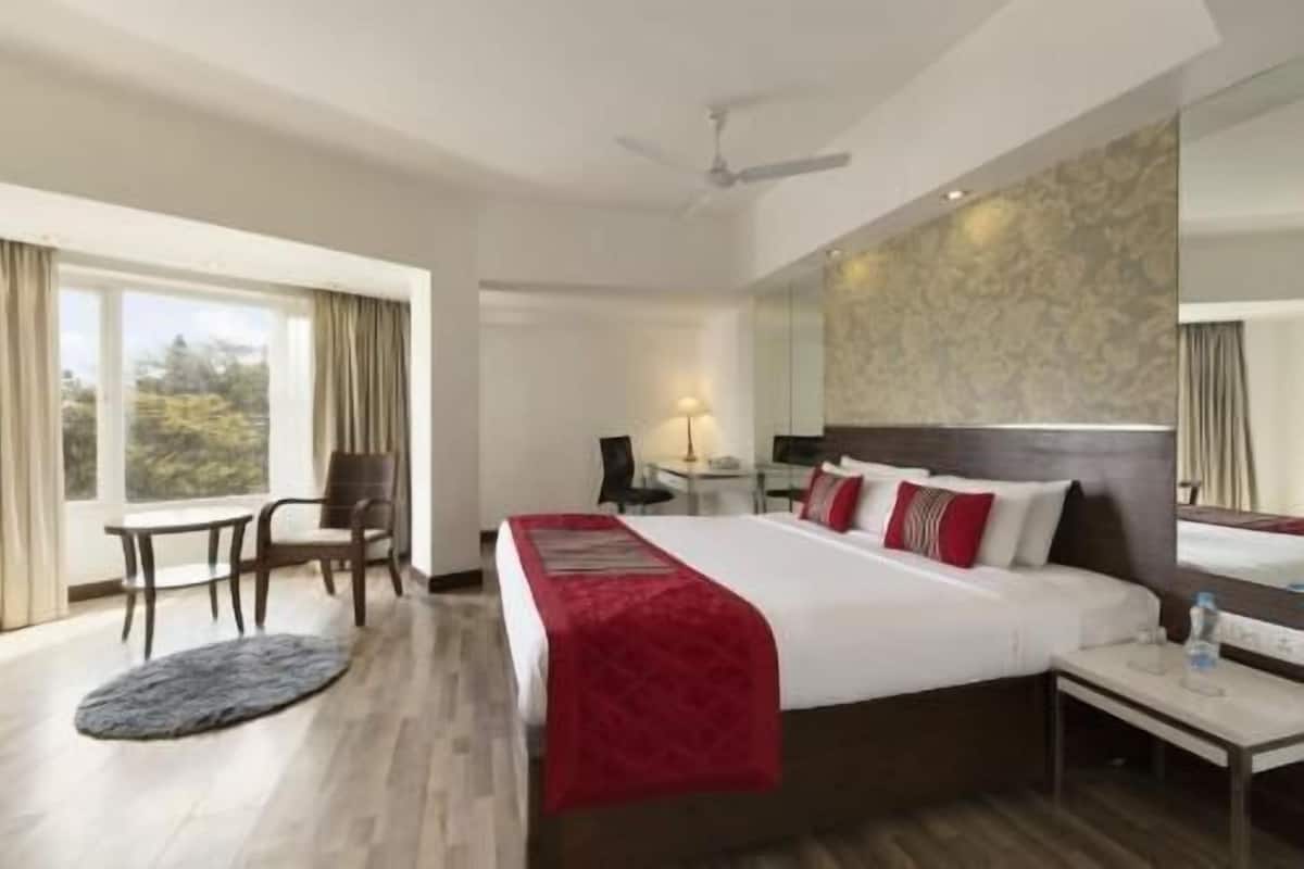 Habitación de hotel con cama, escritorio, silla, mesita, lámpara y ventana con cortinas.