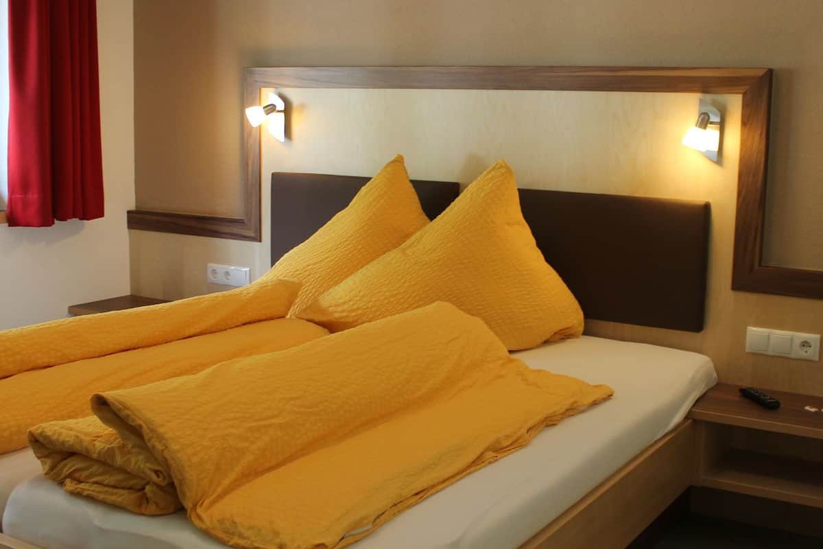 Una cama bien hecha con cabecera, una mesita de noche con control remoto y una ventana con cortinas rojas.