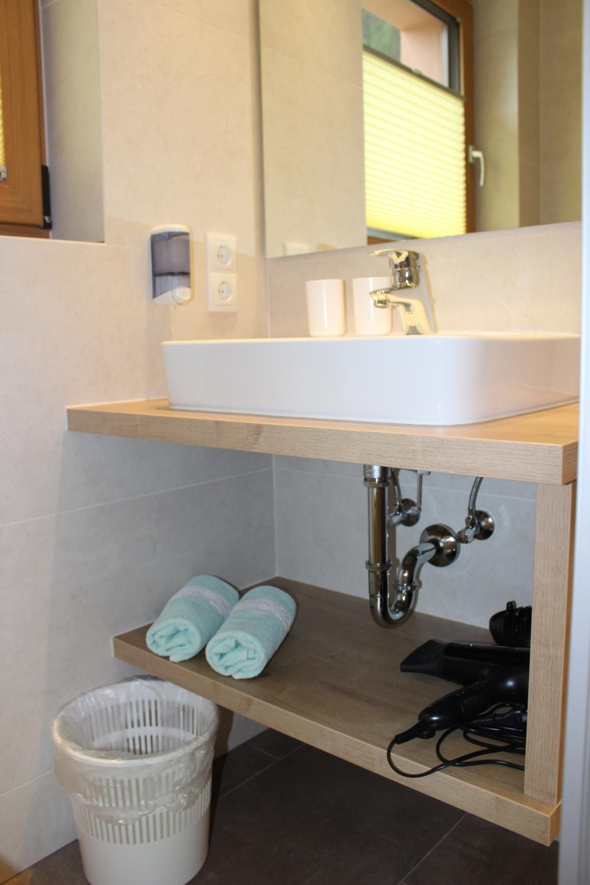 Une salle de bain avec un lavabo blanc, un plan de toilette en bois et un miroir au-dessus. On y trouve deux serviettes sur une étagère et une poubelle blanche.