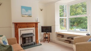 A living room with a fireplace, a television on a stand, a window with a view of greenery, and a couch. - House, Attractive Views Of The Byes, Family and Pet Friendly (Sidmouth)