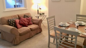 A living room with a beige sofa, a dining table set for four, and a window with a view of a building. - House, Attractive Views Of The Byes, Family and Pet Friendly (Sidmouth)
