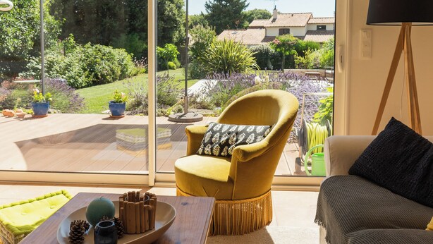 A room with a large sliding glass door leading to a garden, a yellow armchair, a wooden table with a decorative plate, and a black floor lamp.