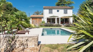 A white house with a swimming pool, surrounded by greenery and a wooden deck.