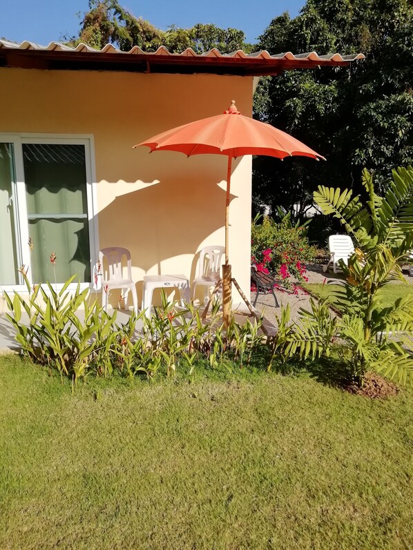 Una casa con techo de tejas, puerta corrediza de vidrio y un pequeño patio con una sombrilla roja, sillas de plástico blanco y plantas en macetas.