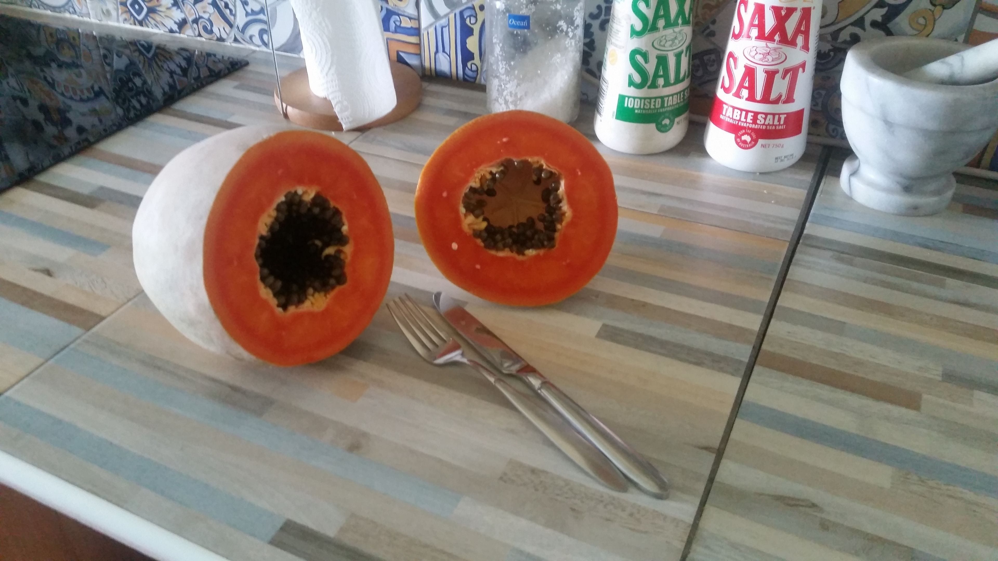 Una encimera de cocina con un papaya cortado por la mitad, un cuchillo y dos botellas de Saka Salt.