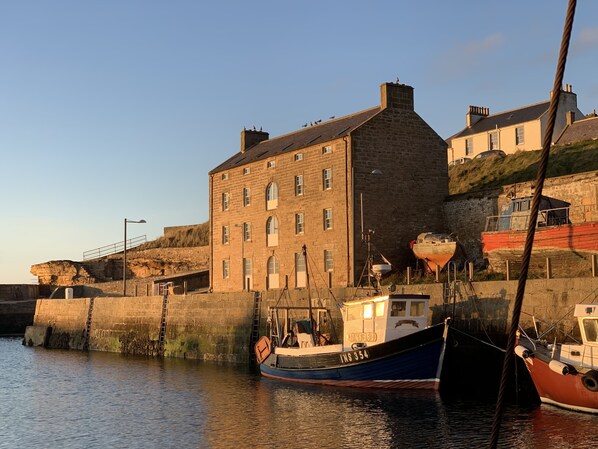 Un bâtiment en pierre doté de nombreuses fenêtres, un quai avec des bateaux, et un ciel dégagé.