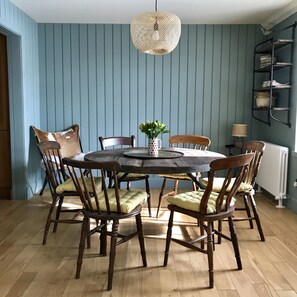 Une salle à manger avec une table ronde en bois entourée de six chaises en bois dotées d’assises rembourrées, un luminaire suspendu en fibres tressées et un mur à rayures bleues.