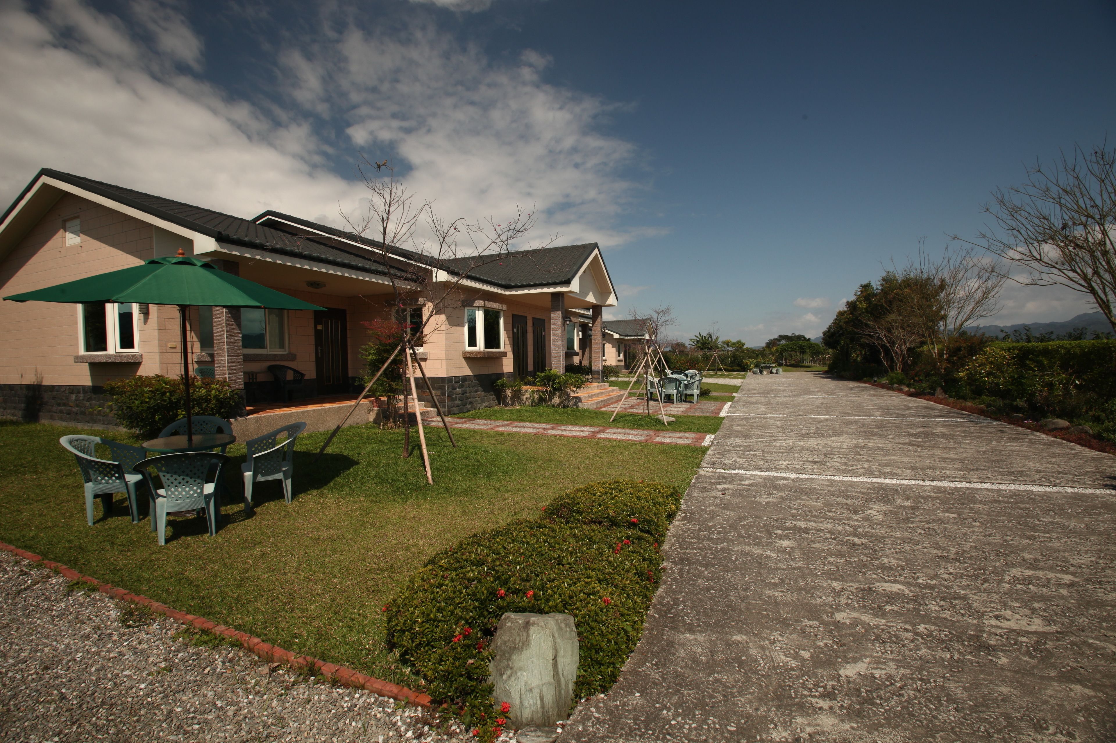 A residential building with a paved driveway, outdoor seating area with chairs and a table, and a green umbrella.